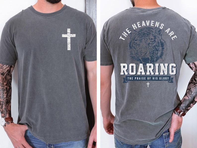 a man wearing a gray t-shirt with a graphic design on the back. The design features a cross and a tiger, along with the text "The heavens are roaring" and "The praise of his glory".