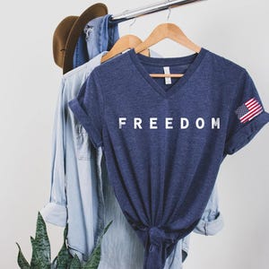a blue t-shirt with the word "FREEDOM" printed on it, hanging on a clothing rack. The t-shirt is accompanied by a denim shirt and a hat, suggesting a casual, laid-back style.