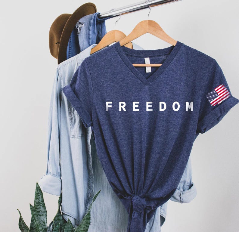 a blue t-shirt with the word "FREEDOM" printed on it, hanging on a clothing rack. The t-shirt is accompanied by a denim shirt and a hat, suggesting a casual, laid-back style.