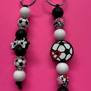 May include: Two keyrings with soccer ball-themed beads. One keyring features a black and white bow, while the other has a soccer ball with a red heart. The keyrings have silver-coloured rings and are set against a pink background.