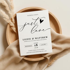 May include: A white wedding invitation with black script that reads "Nothing Fancy, Just Love." The invitation includes the names Lizzie & Matthew, date, time, and location. The invitation is on a wooden plate with decorative elements.