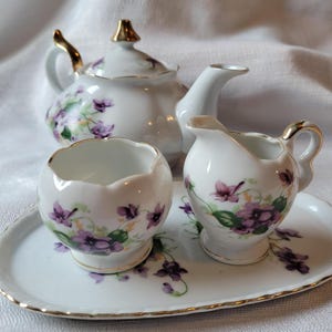 May include: A white porcelain tea set featuring a teapot, creamer, sugar bowl, and serving tray. The set is decorated with purple floral designs and gold trim. The teapot has a gold handle and lid finial. The tray, creamer, and sugar bowl also have gold trim.