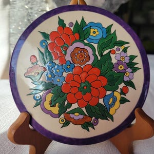 May include: A round, decorative plate with a floral design. The plate features a cream-colored center with a vibrant bouquet of red, blue, yellow, and purple flowers. The rim is a deep purple, and the plate is displayed on a wooden stand.