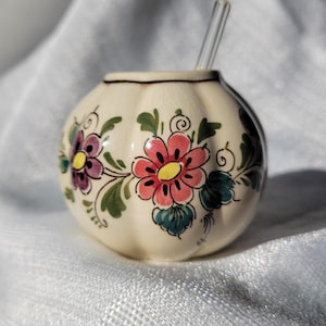 May include: A small, round, cream-colored ceramic container with a clear glass stirring rod. The container is decorated with hand-painted pink, purple, and blue flowers with green leaves. The rim is outlined in black.