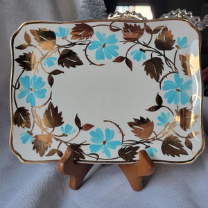 May include: Rectangular white ceramic serving tray with a gold rim and floral design. The tray features light blue flowers, gold and brown leaves, and a gold border. The tray is displayed on a wooden stand.