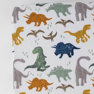 May include: White fitted crib sheet with a dinosaur print. The dinosaurs are in shades of green, blue, orange, and gray. The sheet has a white background with small green and white leaf designs.