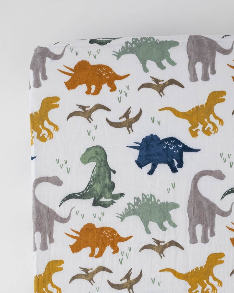 May include: White fitted crib sheet with a dinosaur print. The dinosaurs are in shades of green, blue, orange, and gray. The sheet has a white background with small green and white leaf designs.