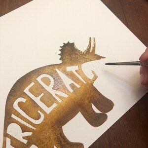 May include: A brown watercolor painting of a triceratops silhouette with the word "TRICERATOPS" written in white inside the silhouette.