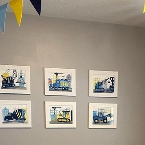 Puede incluir: Seis impresiones enmarcadas de veh&iacute;culos de construcci&oacute;n, incluyendo una grue, un cami&oacute;n volquete, una excavadora y una mezcladora de concreto. Las impresiones est&aacute;n en marcos blancos y est&aacute;n colgadas en una pared gris claro. Una guirnalda amarilla y azul est&aacute; colgada del techo.