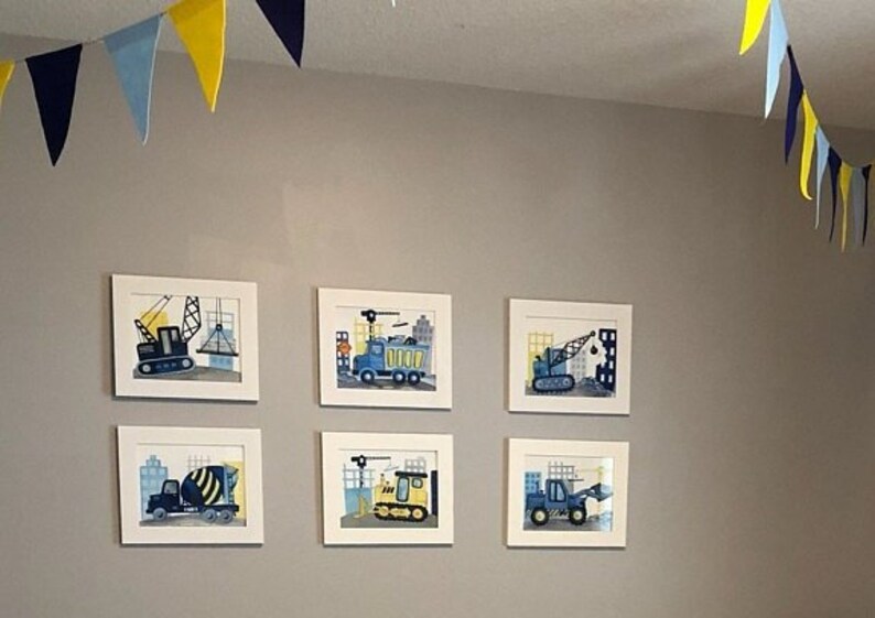 Puede incluir: Seis impresiones enmarcadas de veh&iacute;culos de construcci&oacute;n, incluyendo una grue, un cami&oacute;n volquete, una excavadora y una mezcladora de concreto. Las impresiones est&aacute;n en marcos blancos y est&aacute;n colgadas en una pared gris claro. Una guirnalda amarilla y azul est&aacute; colgada del techo.