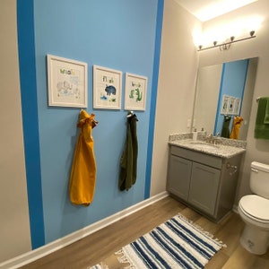 May include: A bathroom with a blue and white wall, a gray vanity with a granite countertop, and a white toilet. There are three framed pictures on the wall with the words "wash", "flush", and "brush" on them. There are two towels hanging on hooks on the wall.