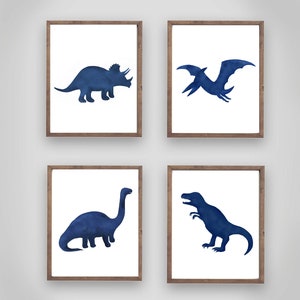 Może przedstawiać: Zestaw czterech oprawionych wydruk&oacute;w przedstawiających sylwetki dinozaur&oacute;w w niebieskiej akwareli. Wydruki obejmują triceratopsa, pterodaktyla, brachiozaura i tyranozaura.