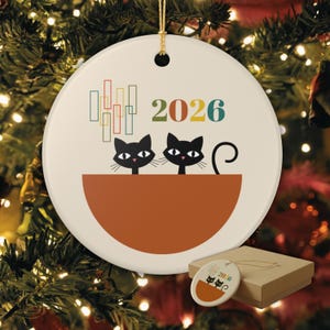 Op de afbeelding: Een rond wit keramieken ornament met twee zwarte katten die uit een grote oranje kom kijken. Boven de katten staat het jaartal 2026 in veelkleurige cijfers naast een reeks verticale rechthoekige vormen in rood, geel en blauwgroen.