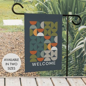 Può includere: Una bandiera da giardino grigio scuro con un motivo geometrico di semicerchi nei toni del verde oliva, arancione bruciato e grigio chiaro. La parola WELCOME è stampata in lettere maiuscole bianche sul fondo. La bandiera pende da un supporto in metallo nero.