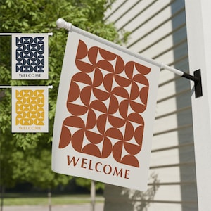 May include: A white outdoor flag with a repeating geometric pattern in rust-orange and white. The word "WELCOME" is printed in rust-orange at the bottom. Two smaller flags with the word "WELCOME" are also visible, one with a navy and cream design, and the other with a yellow and cream design.