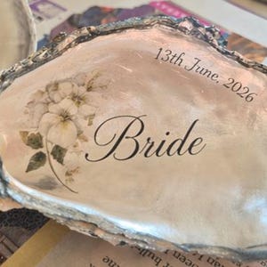 May include: A decorative oyster shell featuring a smooth, pearlescent interior surface. The shell is printed with a delicate floral illustration in soft white and green tones, the word Bride in elegant black script, and the date 13th June, 2026.