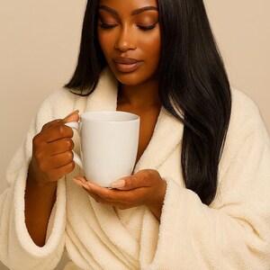 May include: A white ceramic mug held by a person wearing a cream-colored robe. The mug has a simple, cylindrical design. The robe is made of a soft, plush material. The person has long, dark hair and is looking down.