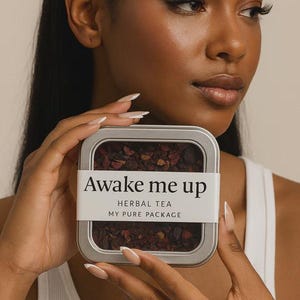 May include: A silver rectangular tin of herbal tea labeled "Awake me up" with the text "Herbal Tea" and "My Pure Package". The tin is held by a person with long nails. The tea inside is a dark reddish-brown color.