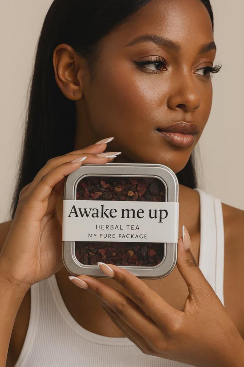 May include: A silver rectangular tin of herbal tea labeled "Awake me up" with the text "Herbal Tea" and "My Pure Package". The tin is held by a person with long nails. The tea inside is a dark reddish-brown color.