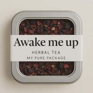 May include: A rectangular, silver-colored tin filled with loose-leaf herbal tea. A white label wraps around the tin, displaying the text "Awake me up" in a bold, black font. Below, the label reads "HERBAL TEA" and "MY PURE PACKAGE". The tea leaves are a mix of dark red and brown.