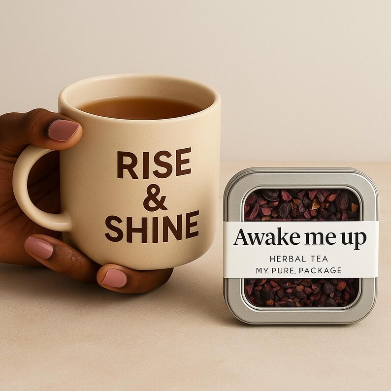 May include: A beige mug filled with tea, featuring the words "RISE & SHINE" in brown lettering. Beside it is a metal tin labeled "Awake me up" with herbal tea. The tea is a dark reddish-brown color.