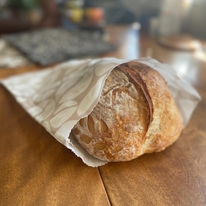 May include: A loaf of freshly baked bread, partially wrapped in a beige paper bag with a leaf pattern. The bread has a golden-brown crust and is resting on a wooden table. The image suggests a bakery or homemade food item.