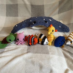 May include: A collection of handmade crochet sea creatures, including a blue manta ray, a green turtle, a pink octopus, and several colorful fish. The toys are arranged on a soft, white and gray checkered blanket.