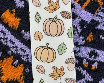 May include: A white bookmark featuring a repeating pattern of pumpkins, leaves, acorns, and pinecones in shades of orange, green, and brown. The bookmark has a glossy finish and is set against a dark blue and purple patterned background.