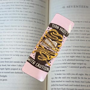 May include: A pink bookmark with the text "TURN PAGES" and "PROCEED WITH CAUTION" in black and yellow. The bookmark features an open book illustration and is resting on the pages of a book.