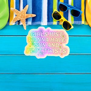 May include: A beach-themed scene with a blue wooden surface. A sticker with the text "Girls Just Wanna Have Sun and Plot Twists" is present. Also, there are blue and yellow sunglasses, a starfish, and a striped towel.