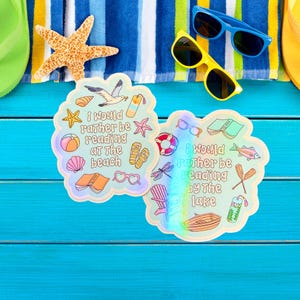 May include: Two die-cut stickers with beach and lake themes, featuring illustrations and the phrases "I would rather be reading at the beach" and "I would rather be reading by the lake." The image also includes sunglasses, a starfish, and a beach towel.