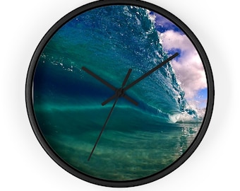 May include: Round wall clock with a black frame. The clock face features a photograph of a breaking ocean wave in shades of blue and green, with a white cloud and blue sky visible. Black clock hands indicate the time.