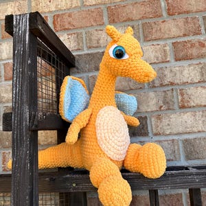 Op de afbeelding: Een handgemaakte, oranje gehaakte drakenknuffel met blauwe vleugels en grote blauwe ogen. De draak heeft een crèmekleurige buik en zit op een houten oppervlak voor een bakstenen muur. De knuffel is gemaakt van zacht, getextureerd garen.