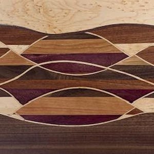 Op de afbeelding: Een rechthoekige houten snijplank met een decoratief golfpatroon. De plank heeft een verscheidenheid aan houttinten, waaronder licht, medium en donkerbruin, met accenten van paars en crème. De plank wordt gepresenteerd op een zwarte metalen standaard.