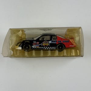 Puede incluir: Un modelo de coche de carreras de fundición a presión en una vitrina de plástico transparente. El coche es negro con detalles en rojo y blanco, con logotipos de patrocinadores y el texto "Coca-Cola 600" y "MAY 28, 2000".