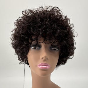 May include: A short, dark brown wig with tight, voluminous curls styled in a rounded shape. The wig is displayed on a smooth, featureless mannequin head with a light brown complexion, pink lips, and dark eyelashes, featuring a white product tag.