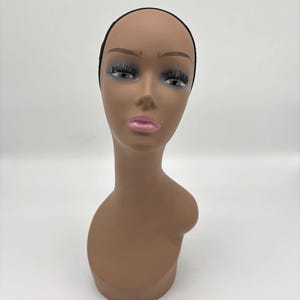 May include: A brown mannequin head with a black cap, painted eyelashes, and pink lips. The mannequin is designed to display wigs and hairpieces. The head is on a white background.