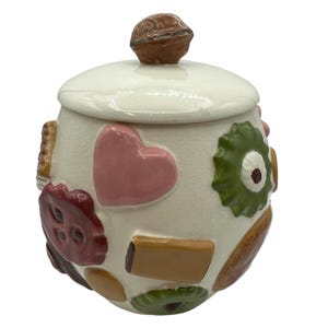 May include: Cream-colored ceramic cookie jar with a lid and a brown knob. The jar is decorated with various colorful shapes, including a pink heart, green flower, and brown rectangles. A charming kitchen accessory.