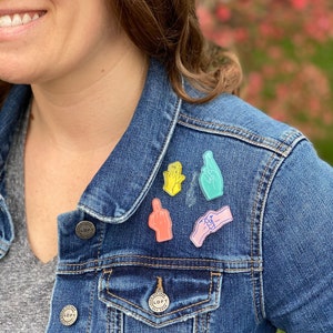 Könnte beinhalten: Eine blaue Jeansjacke mit bunten Emaille-Anstecknadeln, die Handgesten zeigen. Die Anstecknadeln zeigen eine gelbe Hand, eine türkisfarbene Hand mit gekreuzten Fingern, eine rosa Hand, die eine andere Hand hält, und einen pfirsichfarbenen Mittelfinger.