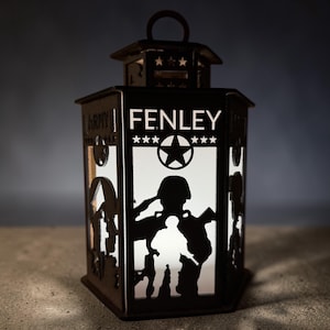 May include: Black metal lantern with a handle, featuring cut-out silhouettes of soldiers and the name "FENLEY" with stars and a military emblem. The word "ARMY" is also visible on the side.