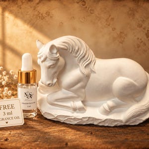 May include: A white ceramic horse figurine in a resting position next to a small clear glass bottle with a gold dropper cap. The bottle label reads NSV Fragrance Oil 3 ml. A white paper tag reads FREE 3 ml FRAGRANCE OIL with a small heart icon.