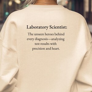 May include: Sand-colored sweatshirt with the text "Laboratory Scientist: The unseen heroes behind every diagnosis—analyzing test results with precision and heart." The word "SAND" is printed at the bottom.