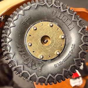 May include: Close-up of a black rubber tire with a gold-colored metal center. The tire has a rugged tread pattern and the word "INJORA" is visible. The tire is marked with "60-20mm-R1.0".