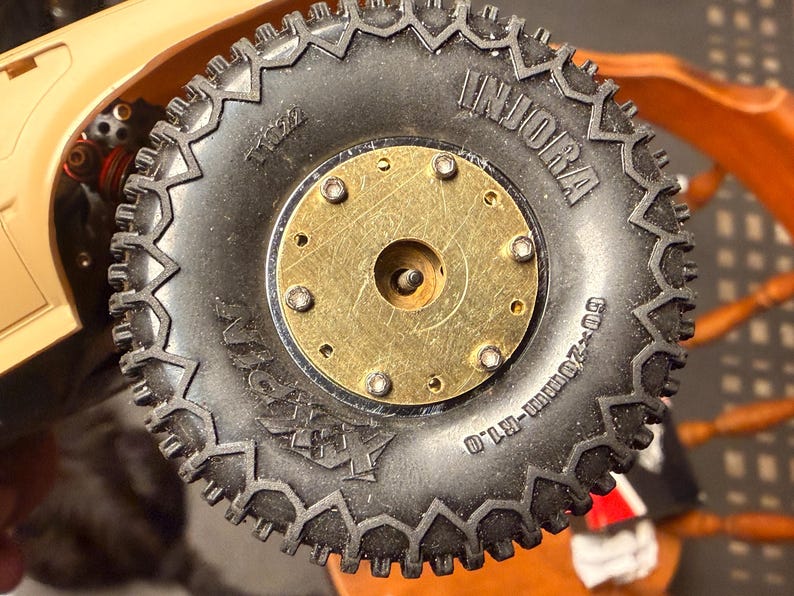 May include: Close-up of a black rubber tire with a gold-colored metal center. The tire has a rugged tread pattern and the word "INJORA" is visible. The tire is marked with "60-20mm-R1.0".