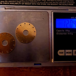 May include: Two small, circular, brass-colored metal pieces with multiple holes are displayed on a digital scale. The scale reads 12.04 grams and has a capacity of 100g with a graduation of 0.01g. The scale is black with a blue digital display.