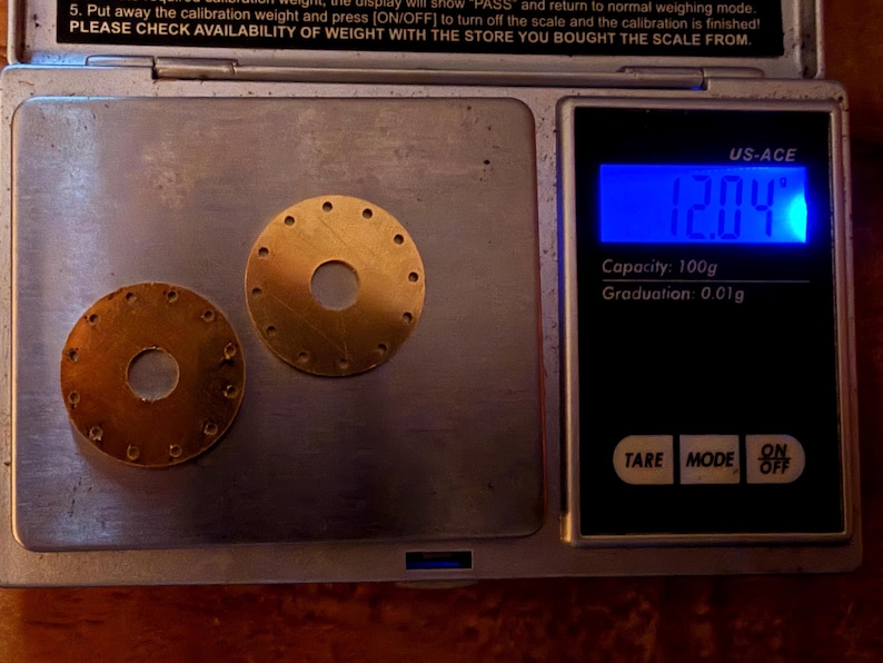 May include: Two small, circular, brass-colored metal pieces with multiple holes are displayed on a digital scale. The scale reads 12.04 grams and has a capacity of 100g with a graduation of 0.01g. The scale is black with a blue digital display.