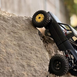 May include: A black remote-controlled pickup truck model climbing a large rock. The vehicle features a matte black body, a roll cage, and off-road tires. The front wheel has a gold-colored rim, while the rear wheels have black rims.