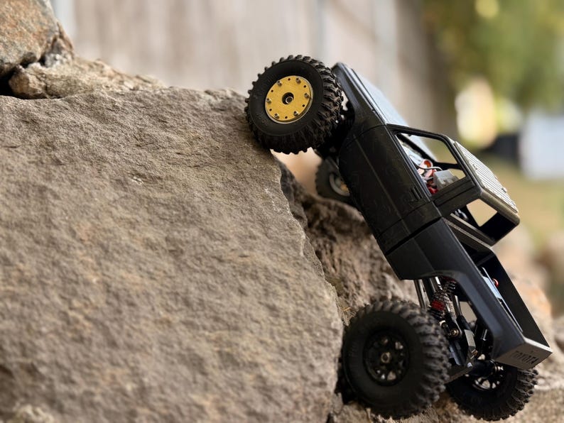 May include: A black remote-controlled pickup truck model climbing a large rock. The vehicle features a matte black body, a roll cage, and off-road tires. The front wheel has a gold-colored rim, while the rear wheels have black rims.