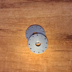 May include: Two circular, flat, grey metal discs, each featuring a central hole and a series of small, evenly spaced perforations arranged in a ring near the outer edge. The discs are stacked slightly offset from one another.