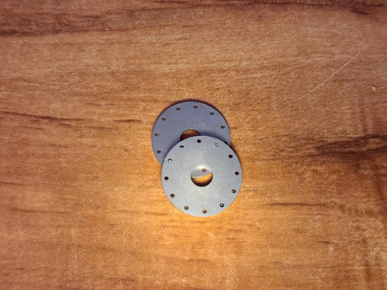 May include: Two circular, flat, grey metal discs, each featuring a central hole and a series of small, evenly spaced perforations arranged in a ring near the outer edge. The discs are stacked slightly offset from one another.